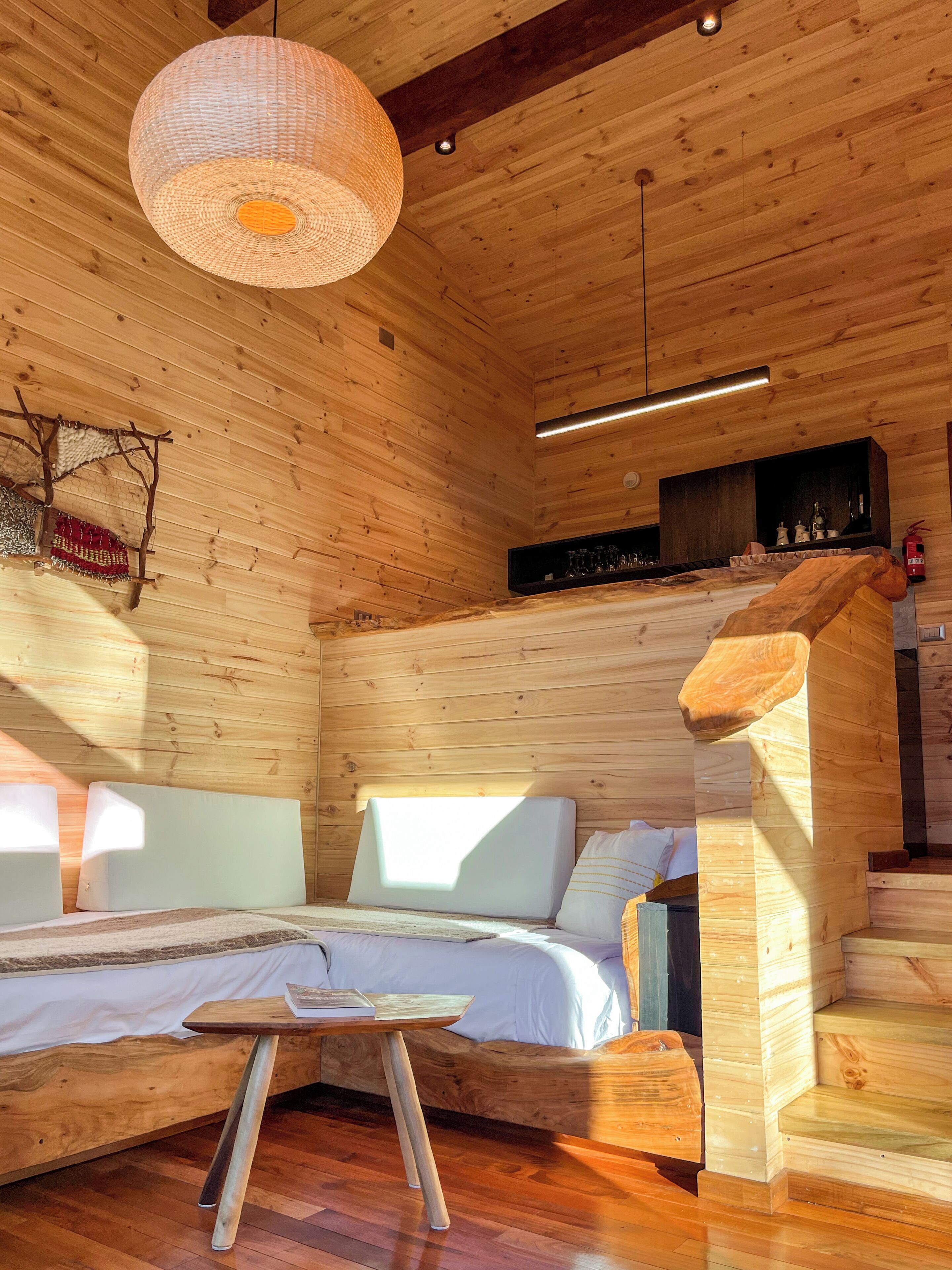 A wooden cabin interior with a bed, a hanging light fixture, and a shelf with decorative items.