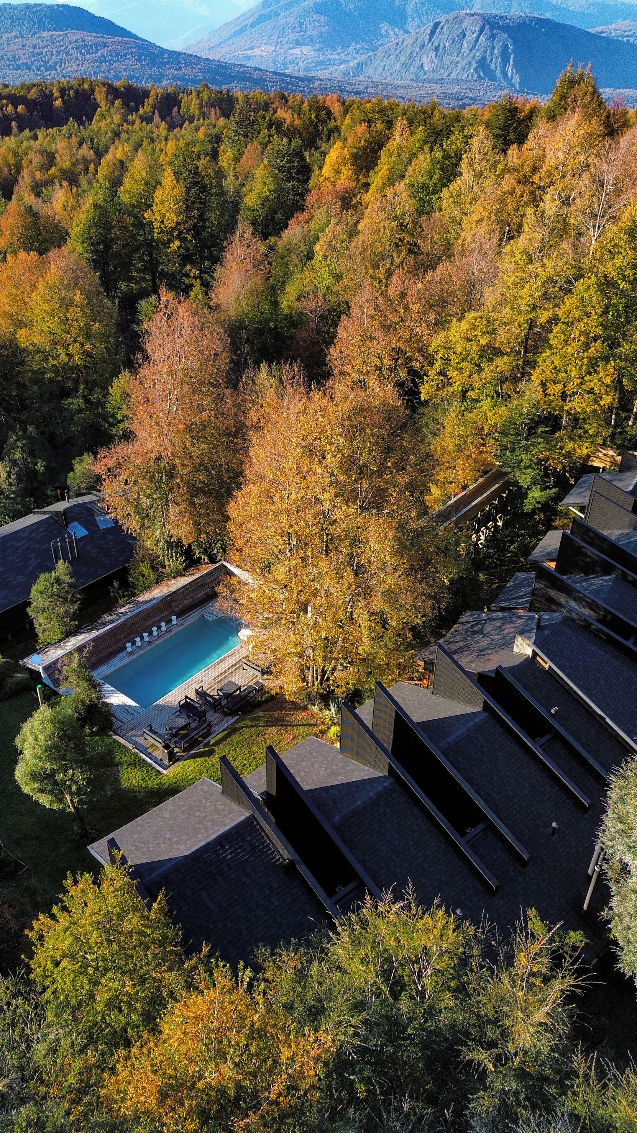 Un complexe hôtelier doté d’une piscine entourée d’arbres aux couleurs automnales, avec une chaîne de montagnes en toile de fond et une architecture moderne.