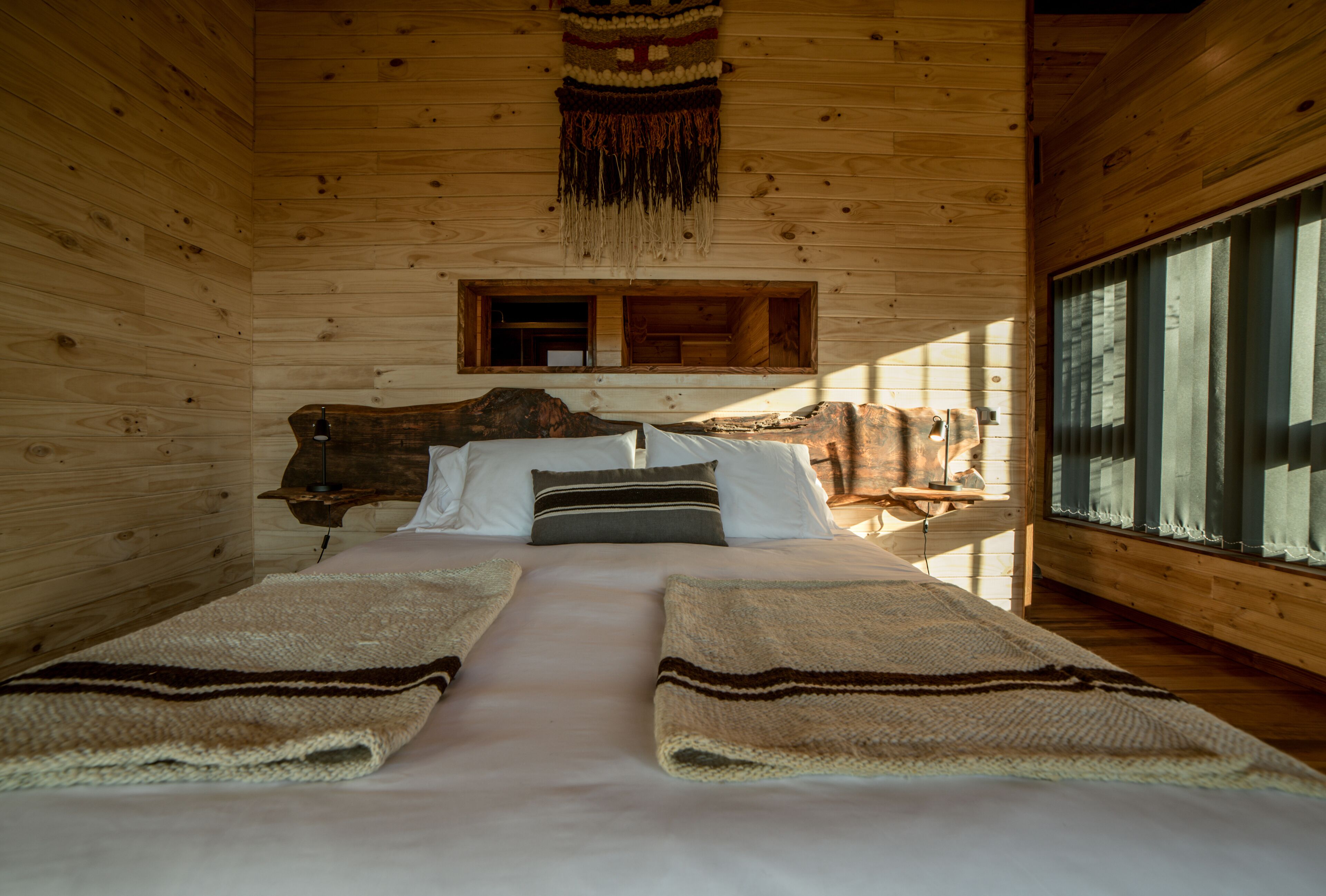 Une chambre dans une cabane en bois, avec un lit, des oreillers, une couverture, une fenêtre et une tapisserie suspendue.