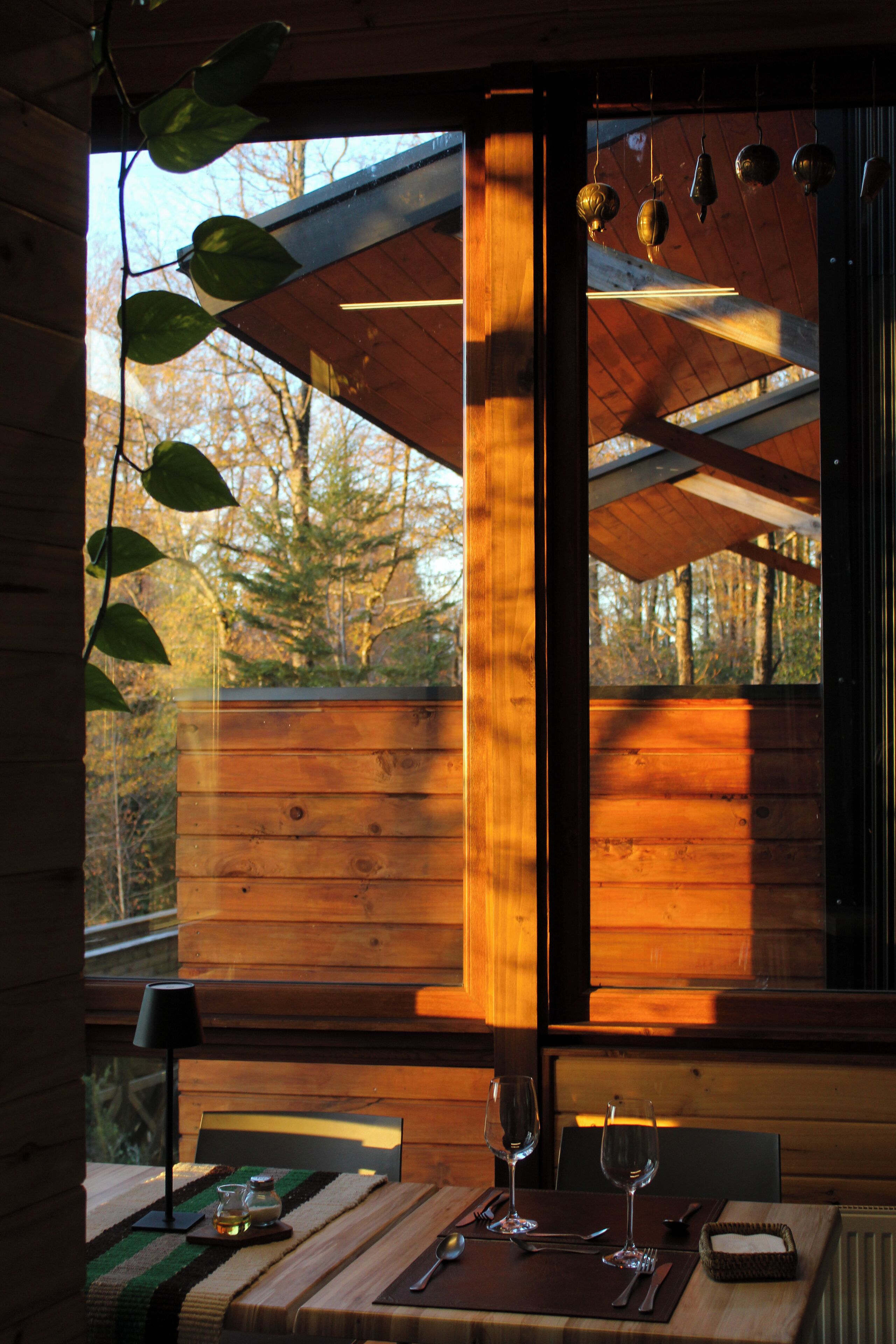 A wooden cabin interior with a dining table set for two, a window with a view of trees, and hanging kitchen utensils.