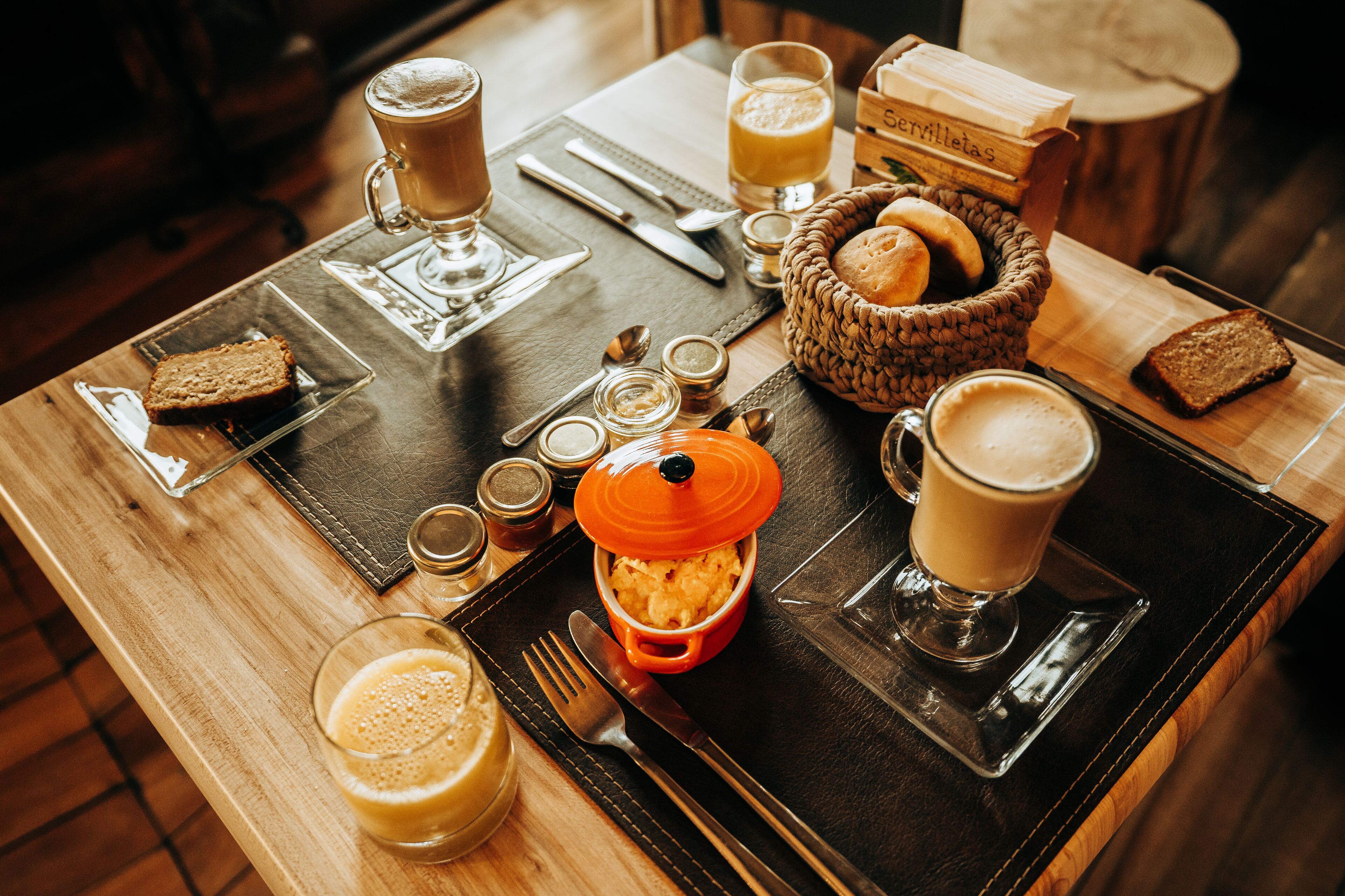 A breakfast setting with a wooden table, coffee cups, bread, and a basket of pastries.