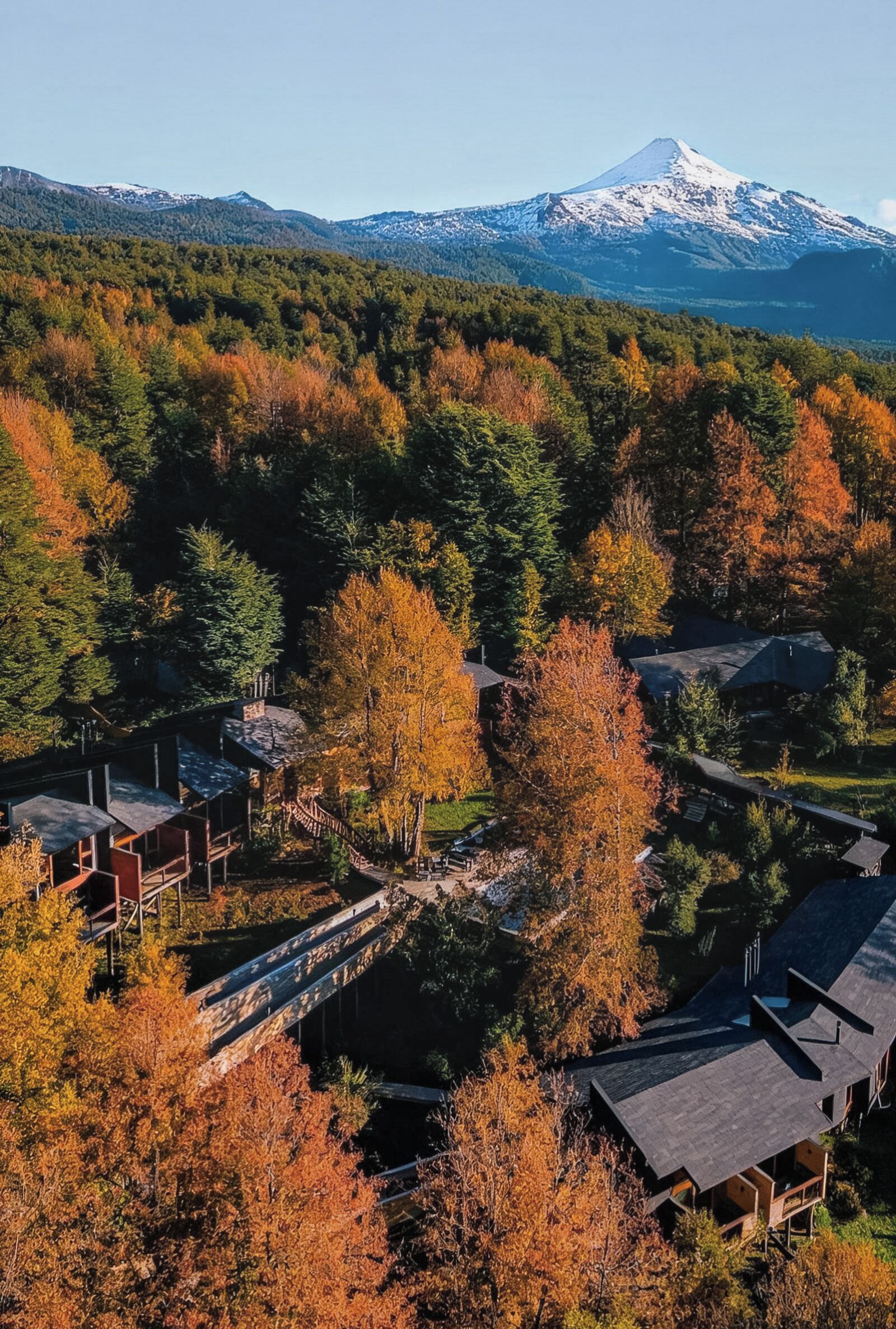 A mountainous landscape with a snow-capped peak, surrounded by autumn-colored trees and a residential area with houses and a wooden bridge.