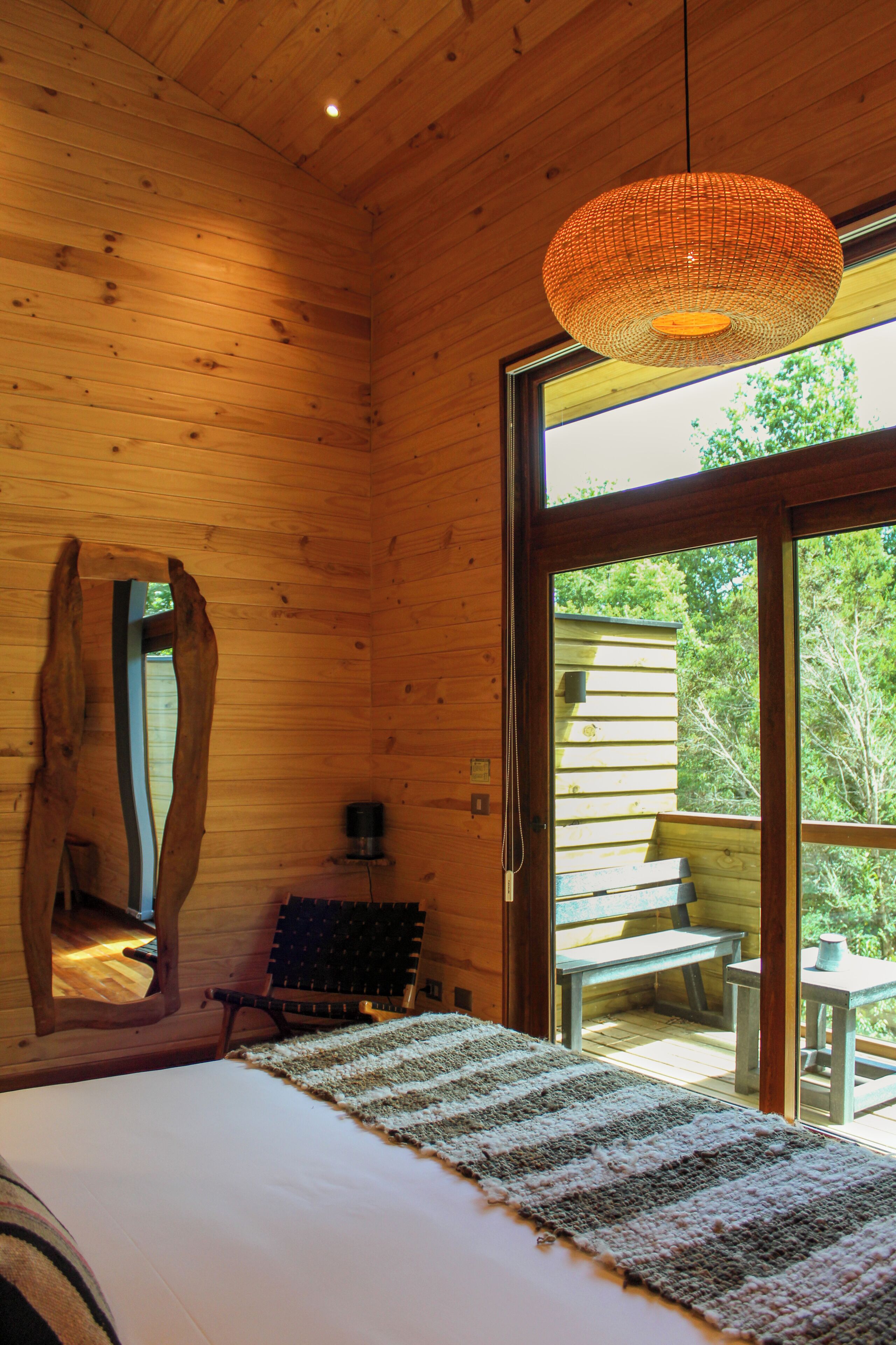 A wooden cabin interior with a bed, a woven light fixture, and a view of the outdoors through a glass door.