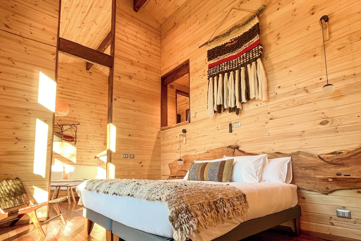 A wooden cabin interior with a bed, wooden floors, a ceiling fan, and a wall with hanging items.