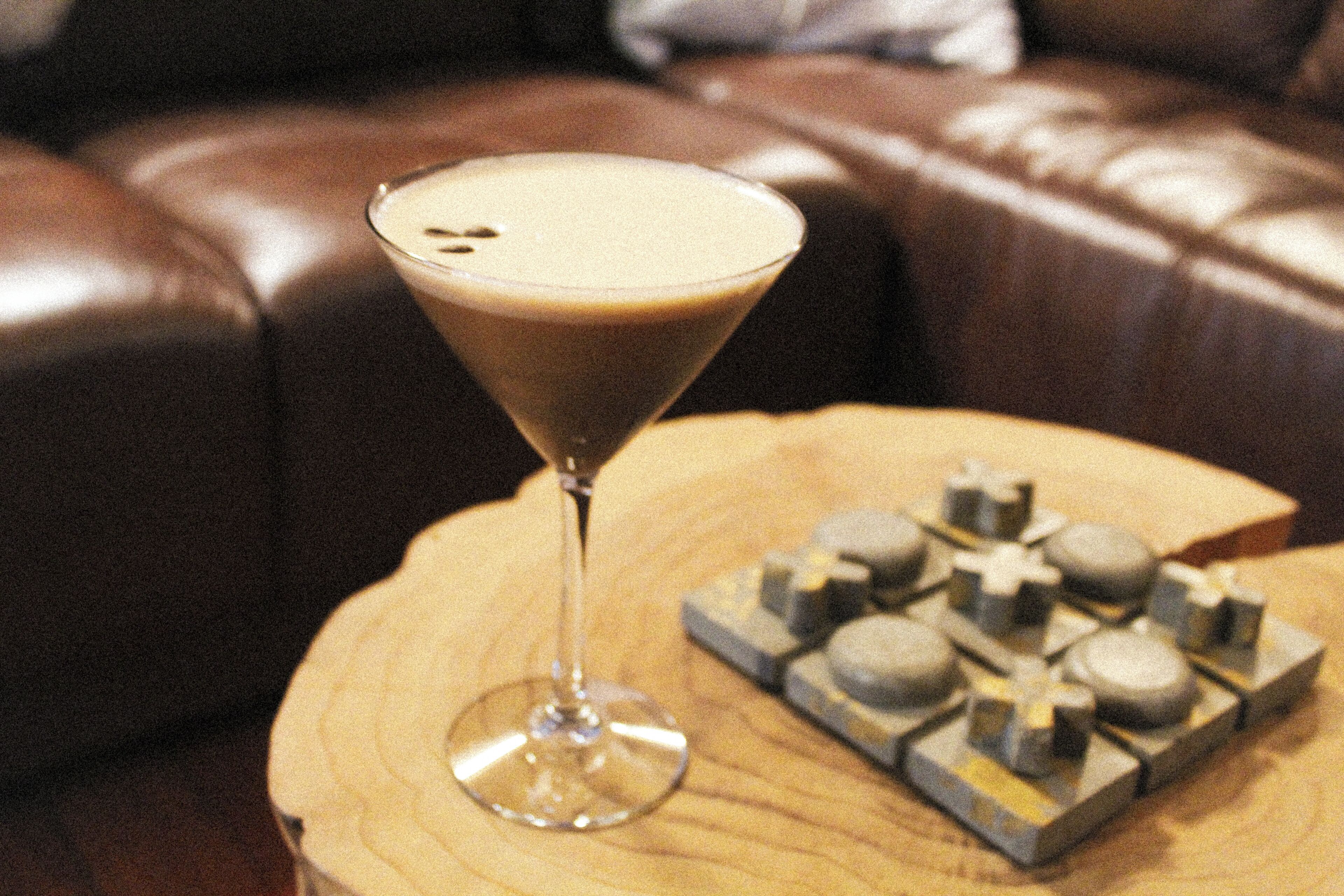 Una copa de martini con una bebida espumosa sobre una mesa de madera, junto a un juego de ajedrez.