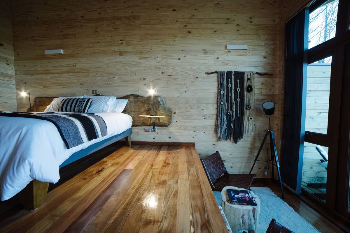 A wooden cabin room with a bed, a desk, and a large window.