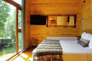 Una habitación de cabaña de madera con una cama, un televisor montado en la pared y un ventanal grande.