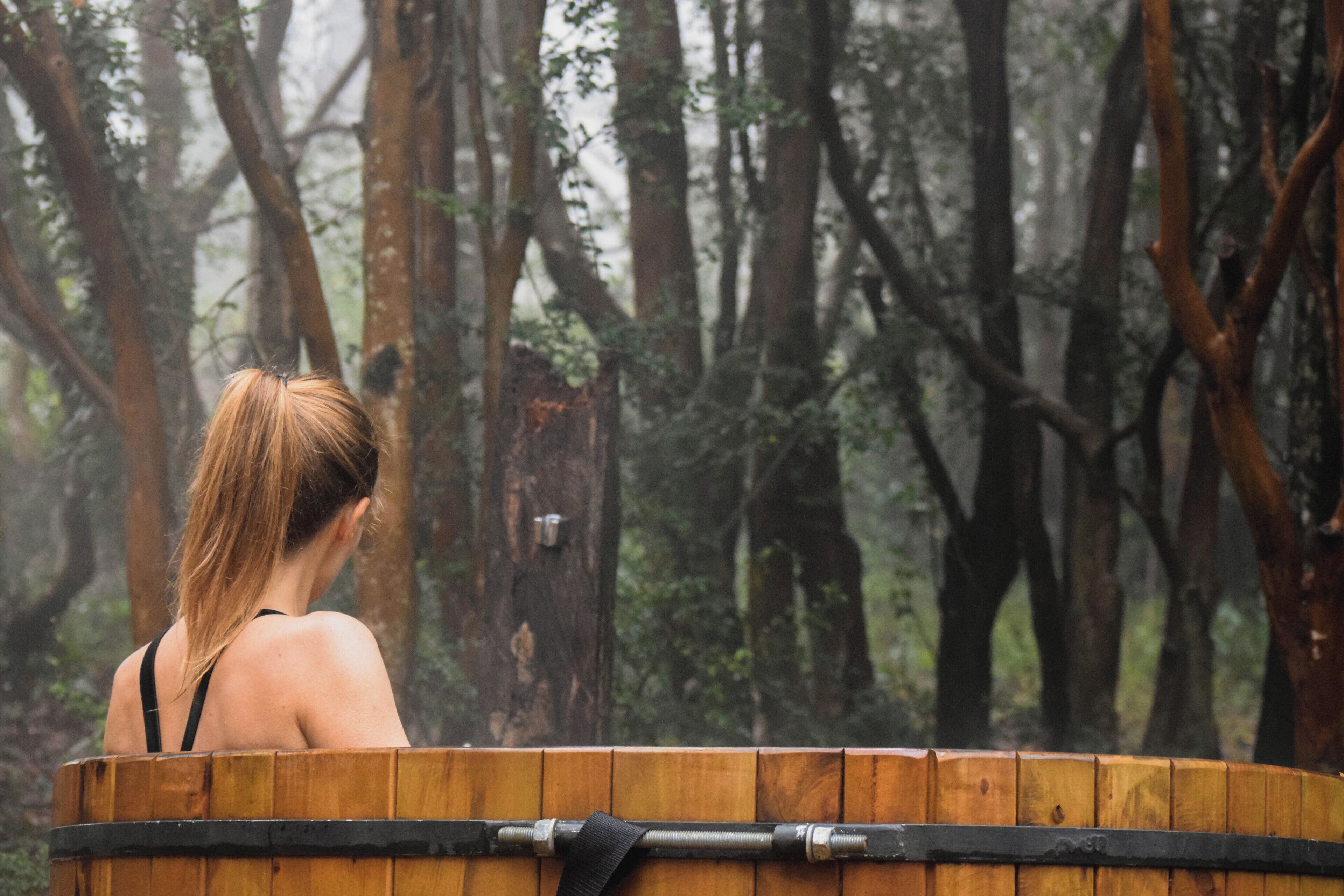 Una persona sentada en un jacuzzi de madera rodeada por un bosque.