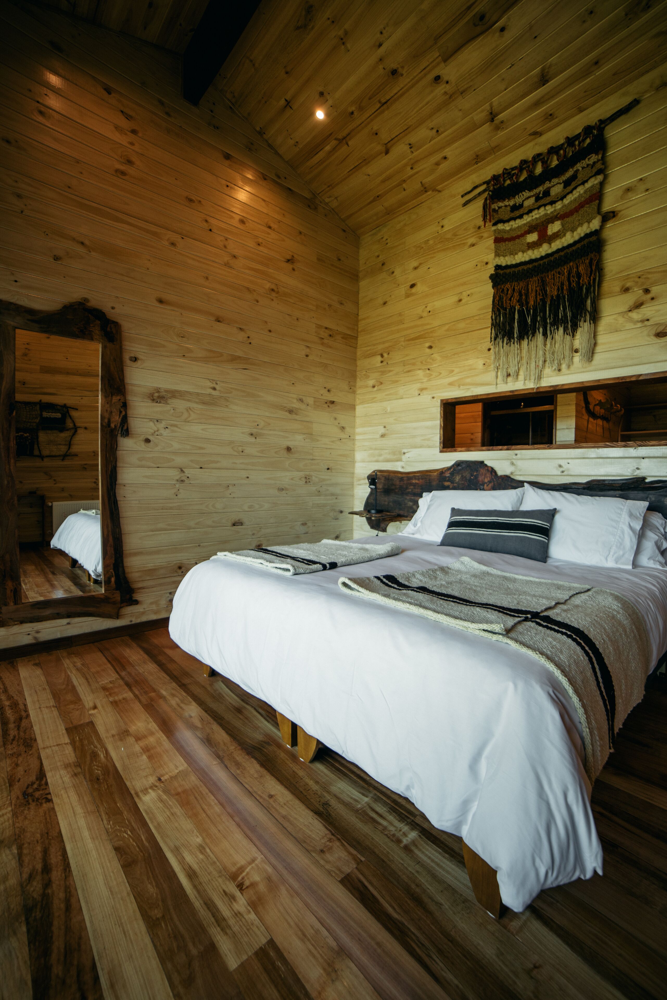 Een houten slaapkamer met een groot bed, houten plafond en een spiegel aan de linkerwand.
