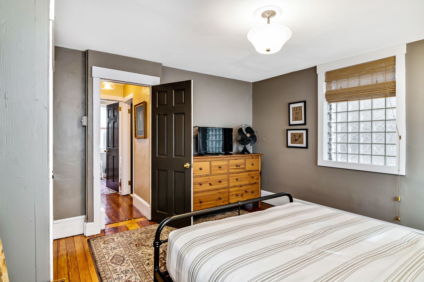 Une chambre à coucher comprenant un lit, une commode, un ventilateur et une fenêtre avec des stores.