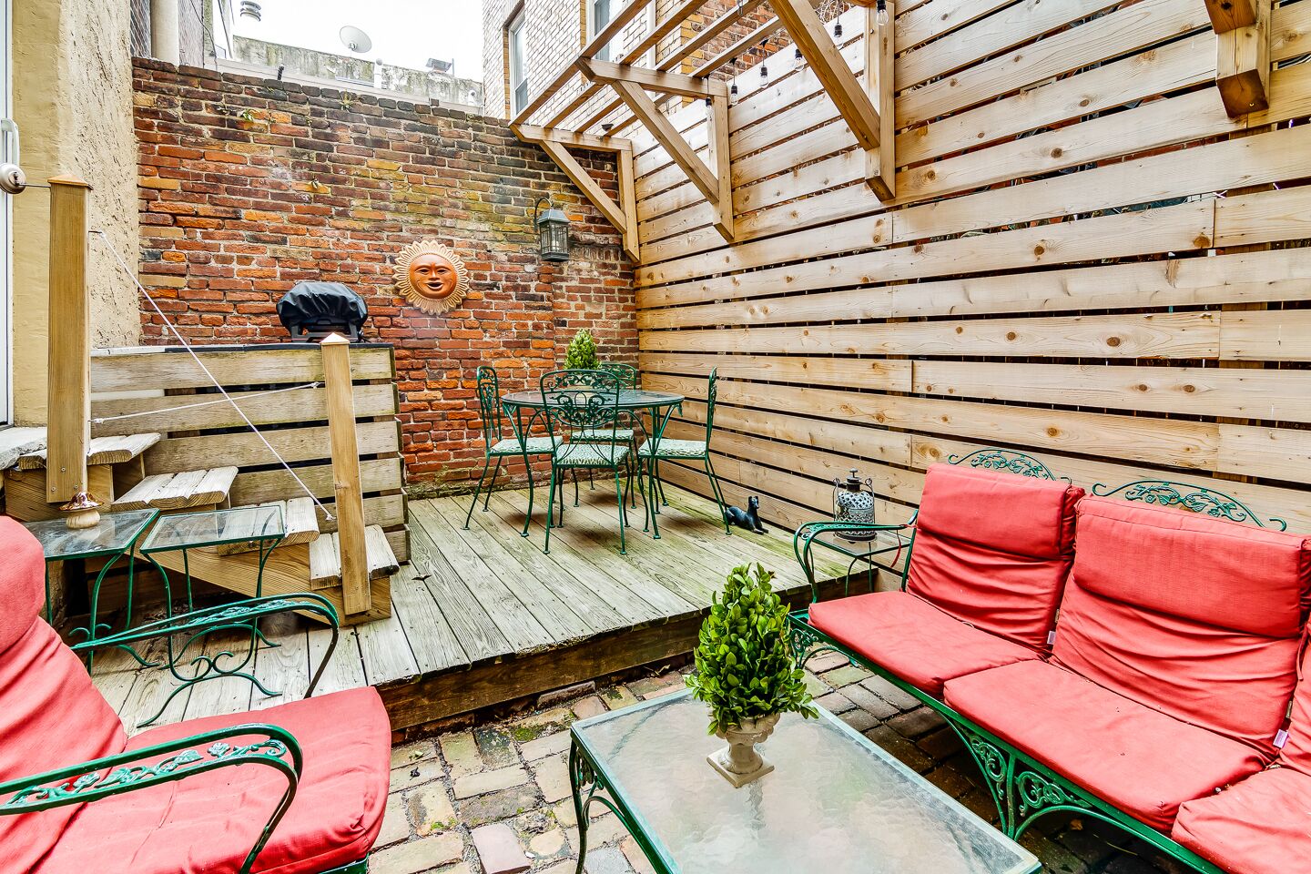 Un patio exterior acogedor con suelo de madera, sillas de metal verde y cojines rojos.
