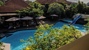 A swimming pool area with lounge chairs and umbrellas, surrounded by trees and buildings with red-tiled roofs. - Marine Bay Sanur (Denpasar)