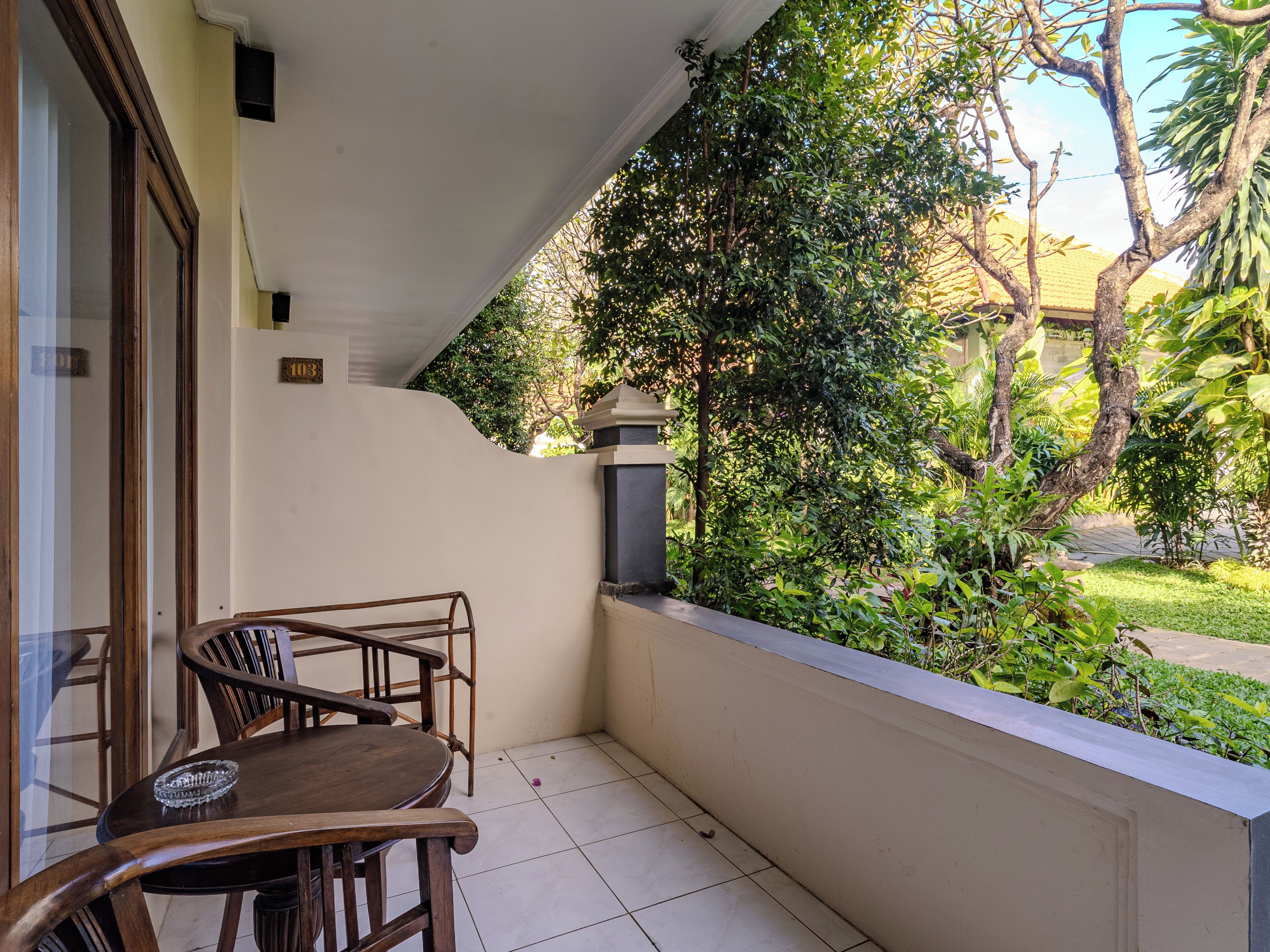 A balcony with a wooden table and chairs, a white wall, and a view of greenery.