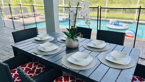 An outdoor dining area with a pool view, featuring a table set with plates and bowls, surrounded by wicker chairs with patterned cushions. - Disney 8BR Villa, Pool + Hot Tub, Theater, Game Room, Themed Bedroom, Sleeps 16+ (Four Corners)