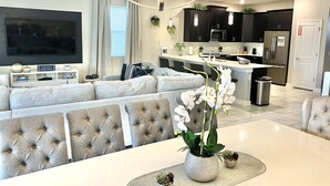 A modern living room with a large flat-screen TV, grey tufted sofa, and a dining table with a vase of white orchids. - Disney 8BR Villa, Pool + Hot Tub, Theater, Game Room, Themed Bedroom, Sleeps 16+ (Four Corners)