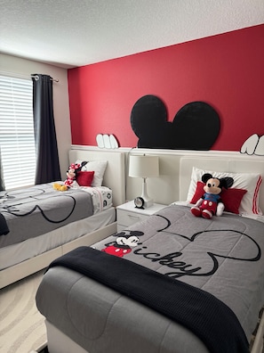 A hotel room with two beds, a Mickey Mouse themed bedspread, a large black Mickey Mouse head decoration on the wall, and a window with curtains. - Disney 8BR Villa, Pool + Hot Tub, Theater, Game Room, Themed Bedroom, Sleeps 16+ (Four Corners)
