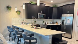 A modern kitchen with dark cabinets, a central island with bar stools, a microwave, a refrigerator, and a sink. - Disney 8BR Villa, Pool + Hot Tub, Theater, Game Room, Themed Bedroom, Sleeps 16+ (Four Corners)