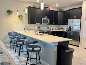 A modern kitchen with dark cabinets, a central island with bar stools, a microwave, a refrigerator, and a sink. - Disney 8BR Villa, Pool + Hot Tub, Theater, Game Room, Themed Bedroom, Sleeps 16+ (Four Corners)