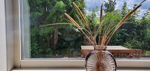 A window with a view of a wooden deck and a glass vase with tall grasses.
