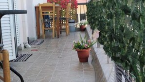 En terrasse med flisegulv, træstole, et bord, plantede planter og en grøn hæk.