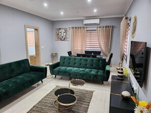 A living room with a green sofa, a coffee table, and a TV. There is a dining table with chairs and a painting on the wall. - Spacious Luxury 3Bed Hse in Tema - Netflix, Wi-fi (Tema)