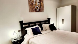 Una cama bien hecha con ropa de cama blanca y dos almohadas oscuras, una mesita de noche negra con una lámpara, y un armario blanco.