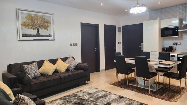 Una sala de estar moderna con un sofá de color oscuro, cojines estampados, una mesa de comedor de vidrio con sillas negras y una lámpara de araña.