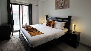 A bedroom with a large bed, a desk, a chair, a nightstand with a lamp, and a painting on the wall. - Elegant & Luxury Gem-5 Star Location -Pools, Gym! (Accra)