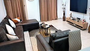 A modern living room with a black leather sofa, a round wall clock, a flat-screen TV, and a wooden entertainment unit. - Elegant & Luxury Gem-5 Star Location -Pools, Gym! (Accra)
