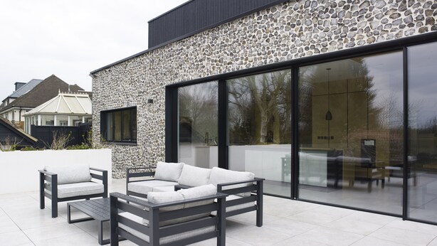 A modern outdoor patio with a stone wall, a large glass sliding door, and a seating area with a sofa and chairs.