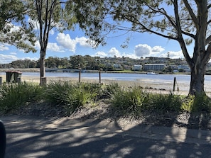 A riverside walkway with trees and grass, a bench, and a view of a river and buildings in the distance.