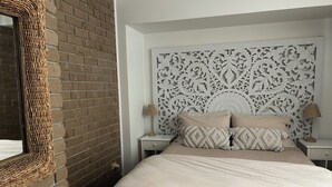 Une chambre avec un mur en briques, une tête de lit décorative, un lit avec une literie beige et deux tables de chevet avec des lampes.