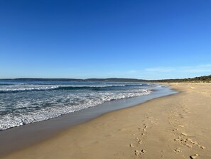 A beach with footprints leading to the water, a clear sky, and a distant shoreline. - location location 5min walk to beach, lake, restaurants and shops (Merimbula)