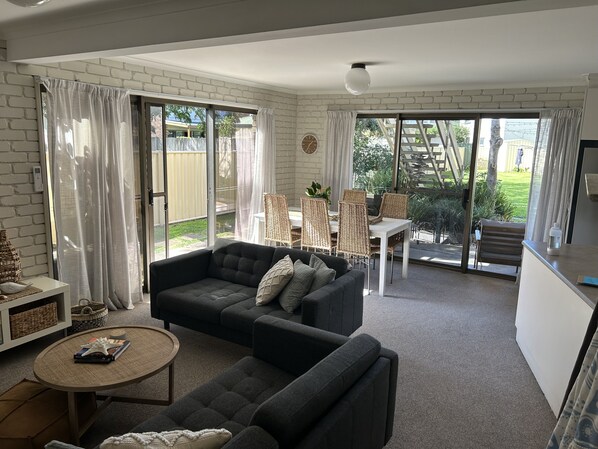 A living room with a dark grey sofa, a round coffee table, and a dining area with wicker chairs. - location location 5min walk to beach, lake, restaurants and shops (Merimbula)