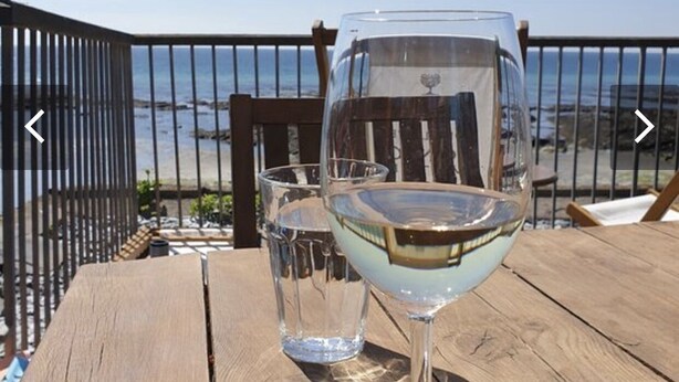 A wooden table with two wine glasses, one filled with white wine, and a clear glass beside it. The table is outdoors with a view of the ocean and a metal railing in the background. - Torpoint - Apartment - Sleeps4 - Seaside - Parking (Torpoint)
