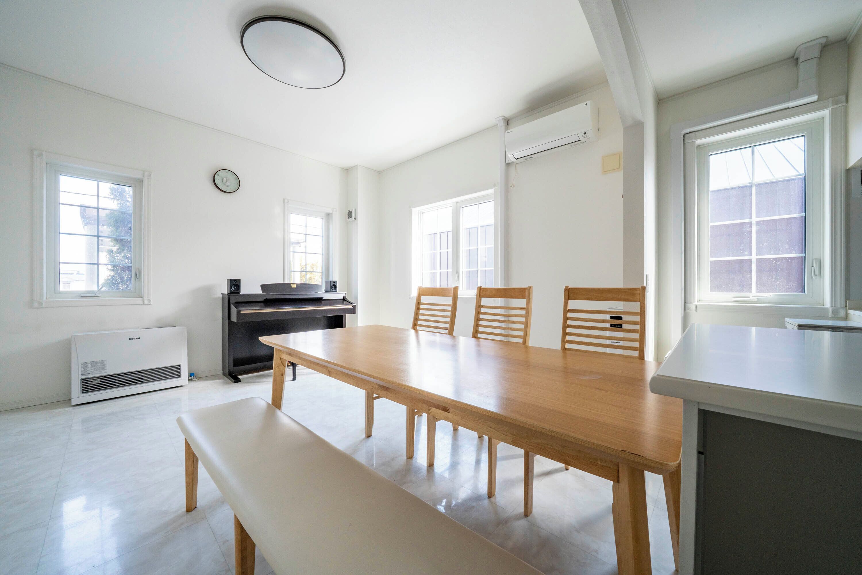 A spacious room with a large wooden dining table, four chairs, a bench, a piano, and a large window providing natural light.