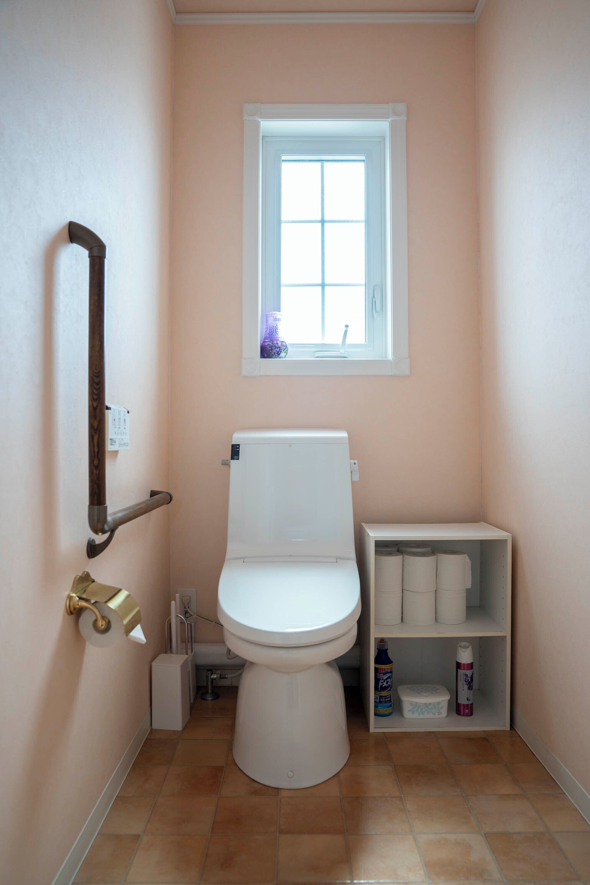 A small bathroom with a toilet, a shelf with toilet paper, and a window above the toilet.