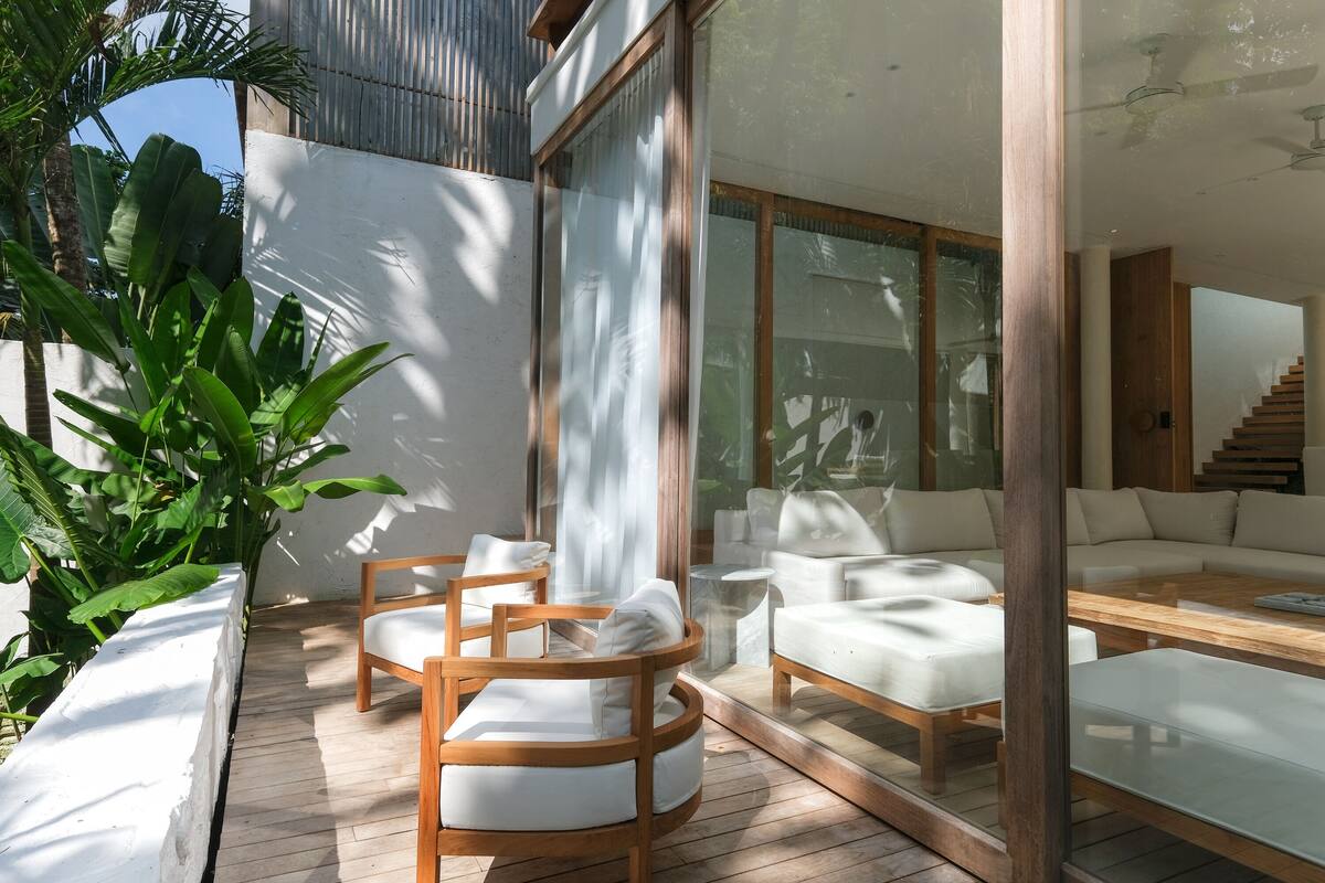 Un espace extérieur moderne, aménagé avec des chaises en bois et des coussins blancs, une cloison en verre et une table en bois.