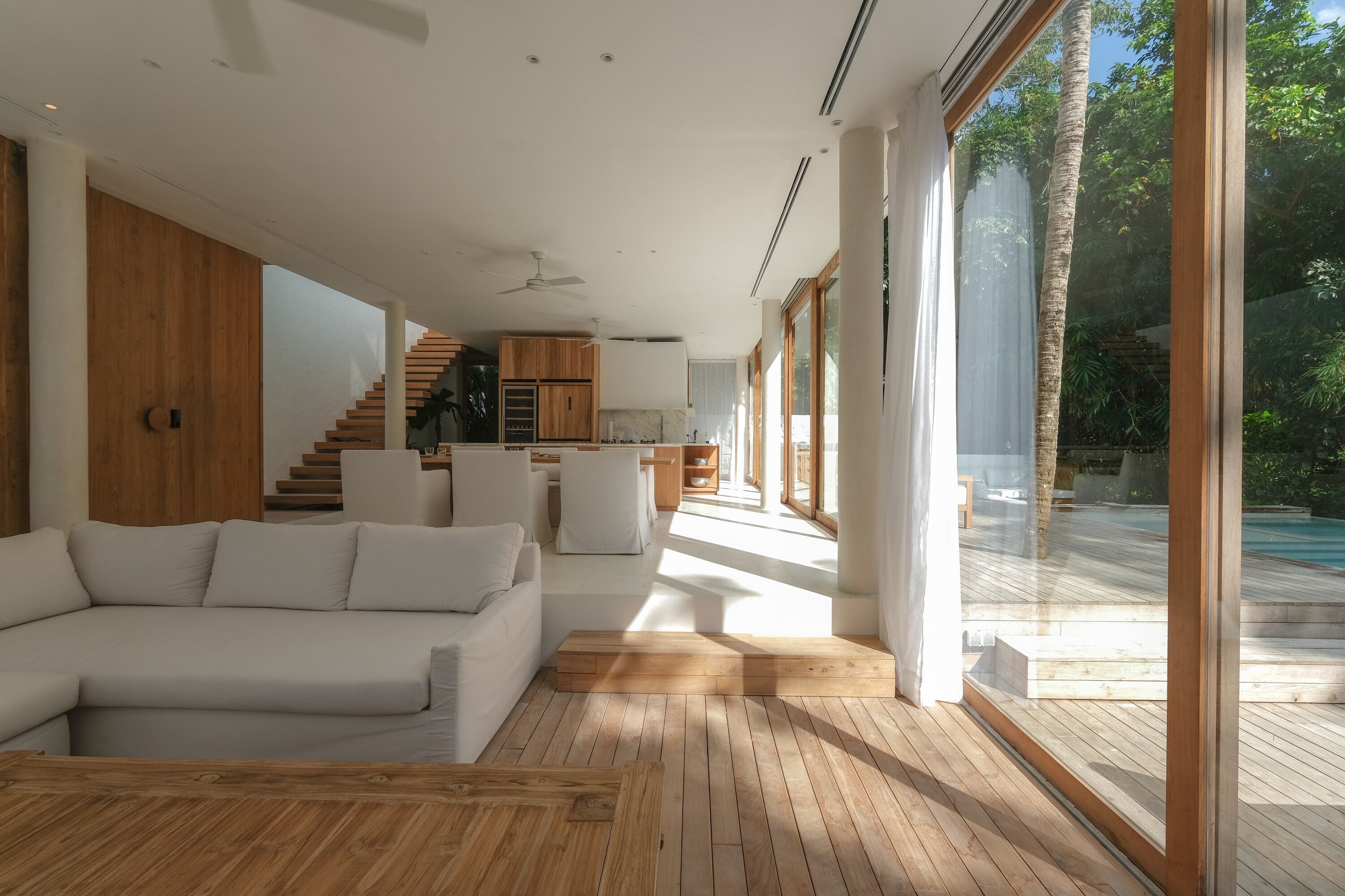 A modern living room with a wooden floor, white sofa, and large windows overlooking a pool.
