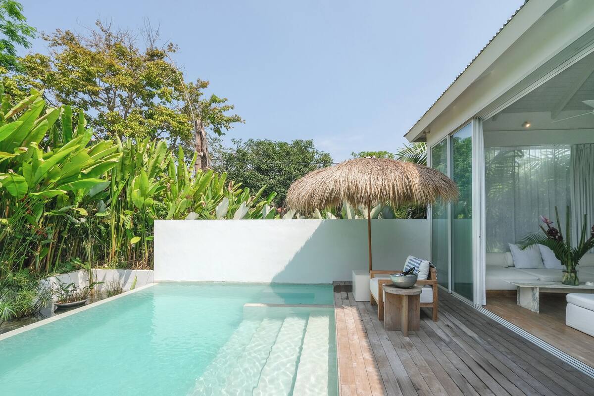 Un espace aménagé au bord de la piscine, avec une terrasse en bois, un parasol en chaume et un coin salon doté de coussins et d’une table.