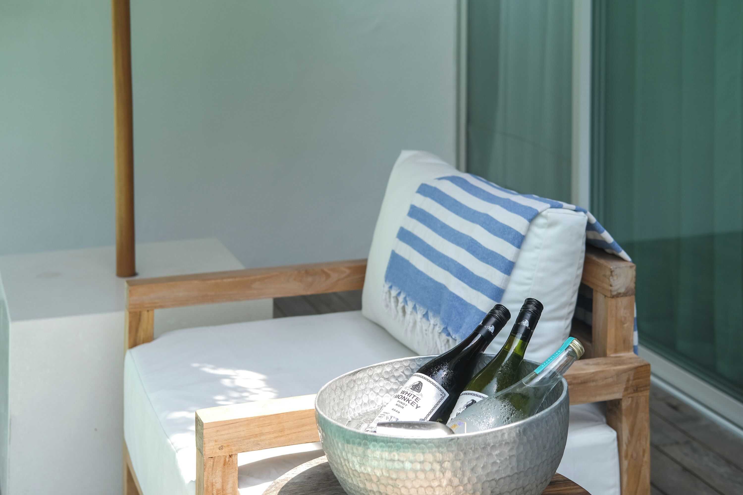 A wooden chair with a white cushion and a blue and white striped pillow. In front of the chair is a glass bowl containing two bottles of wine and a bottle of champagne.