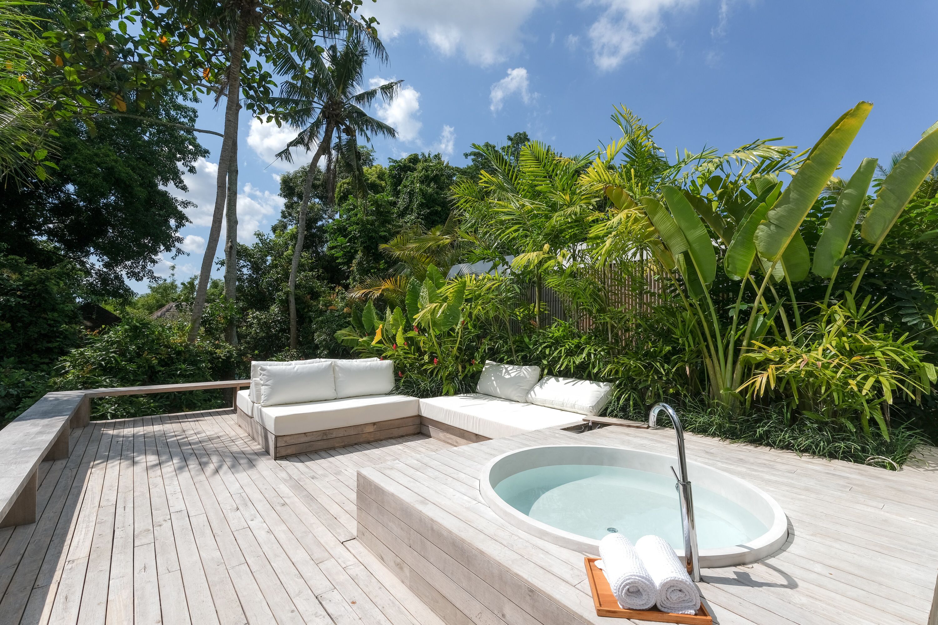 Una terraza de madera con jacuzzi, asientos acolchados blancos y exuberante vegetación tropical.