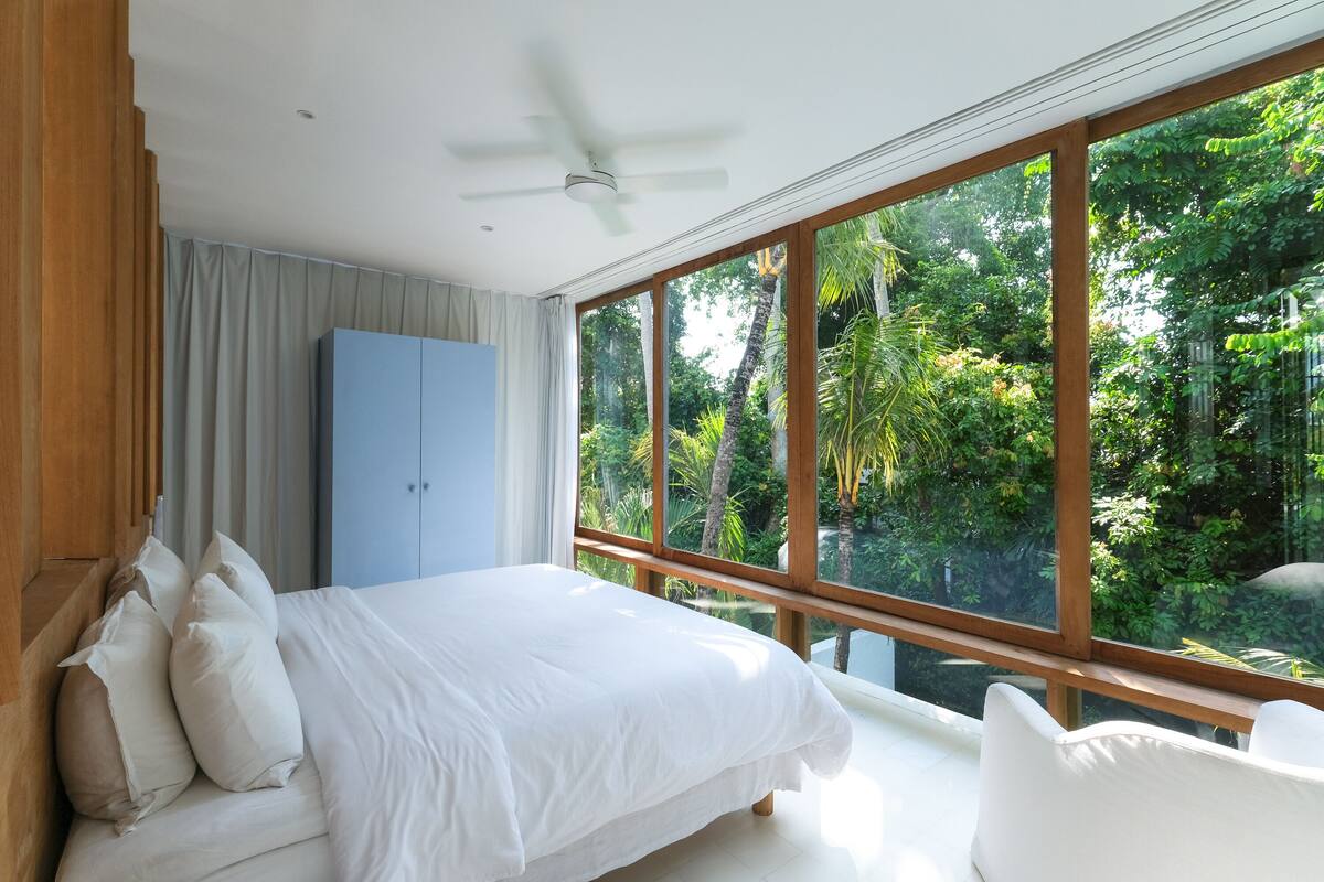 Une chambre avec une grande fenêtre, un lit, une armoire et un ventilateur de plafond.