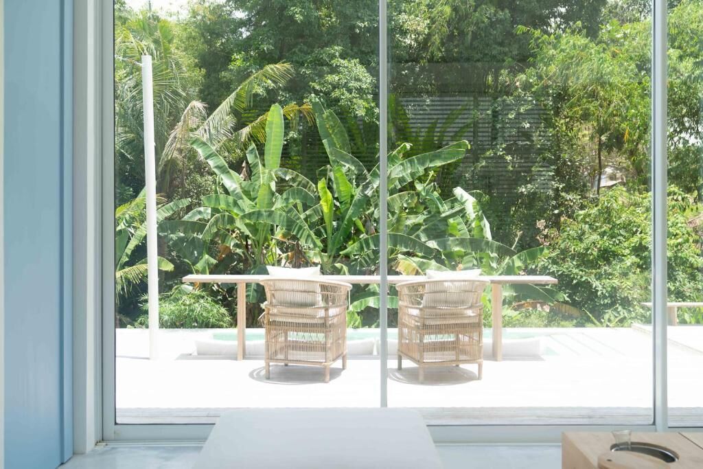 Habitación con un ventanal grande que da a un jardín exuberante con plantas de banano y un muro de ladrillo. Hay una mesa blanca con dos sillas de mimbre.