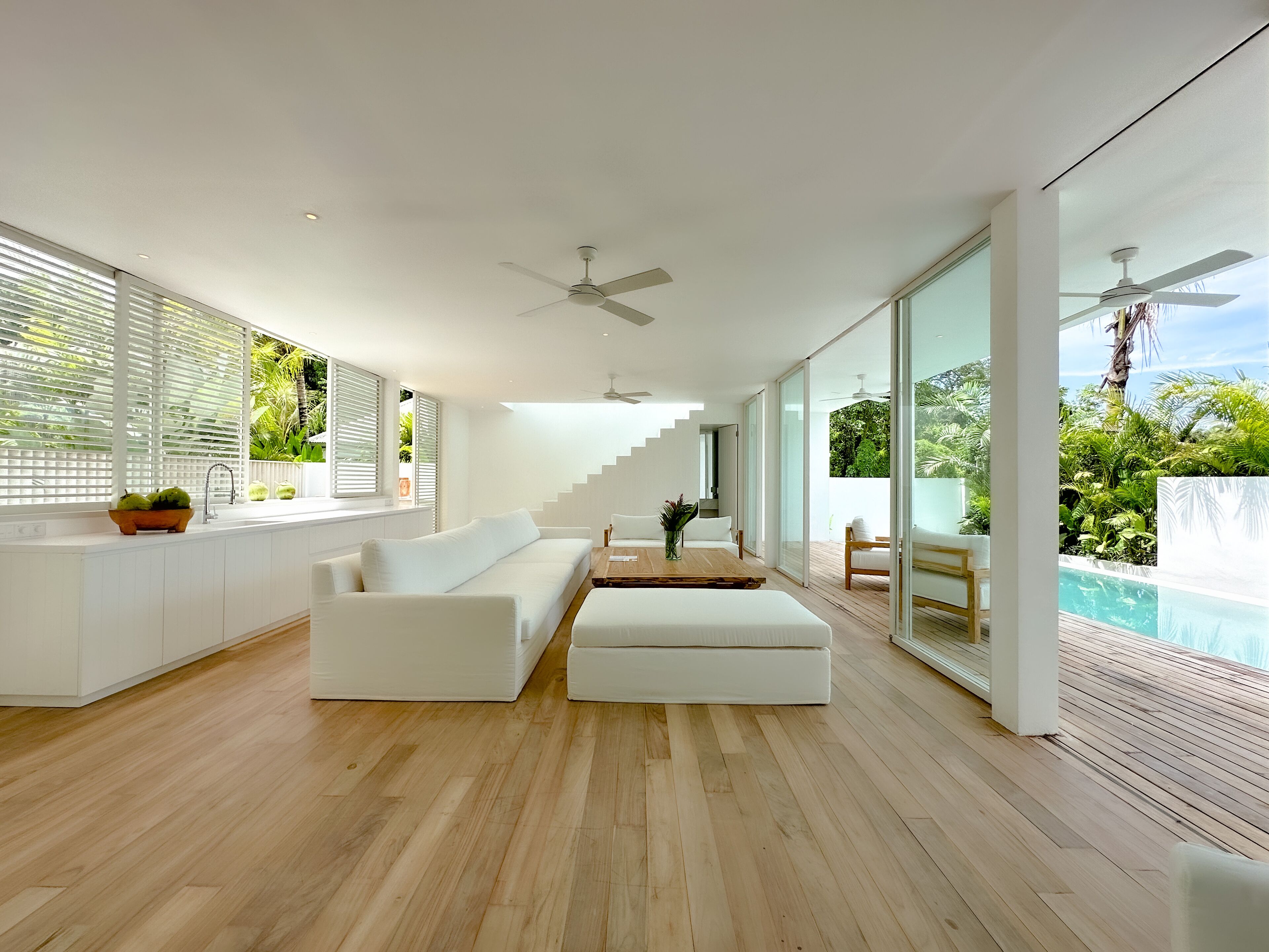 A modern living room with a white sofa, wooden flooring, and large windows overlooking a pool.