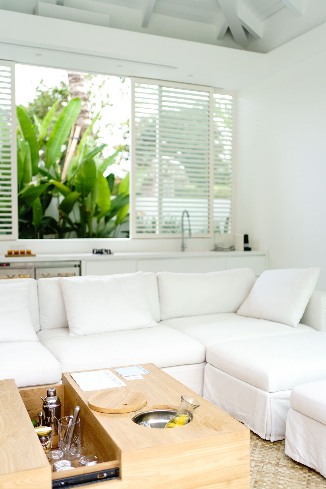 Una sala de estar moderna con un sofá seccional blanco, una mesa de centro de madera con copas de cóctel y un ventanal con persianas.