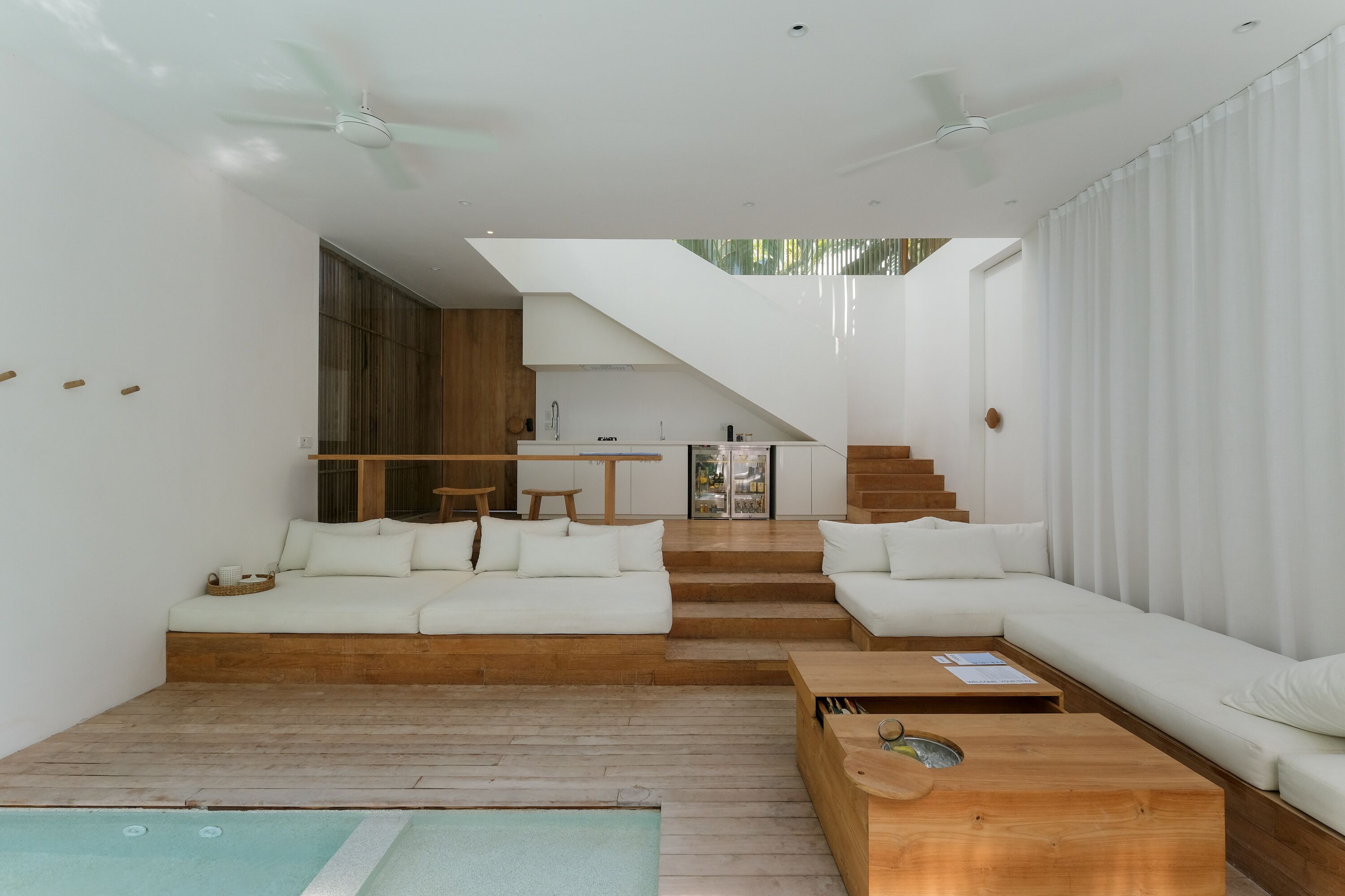Un espacio moderno con sala de estar, piso de madera, sofás blancos, piscina y una escalera que lleva a una cocina.