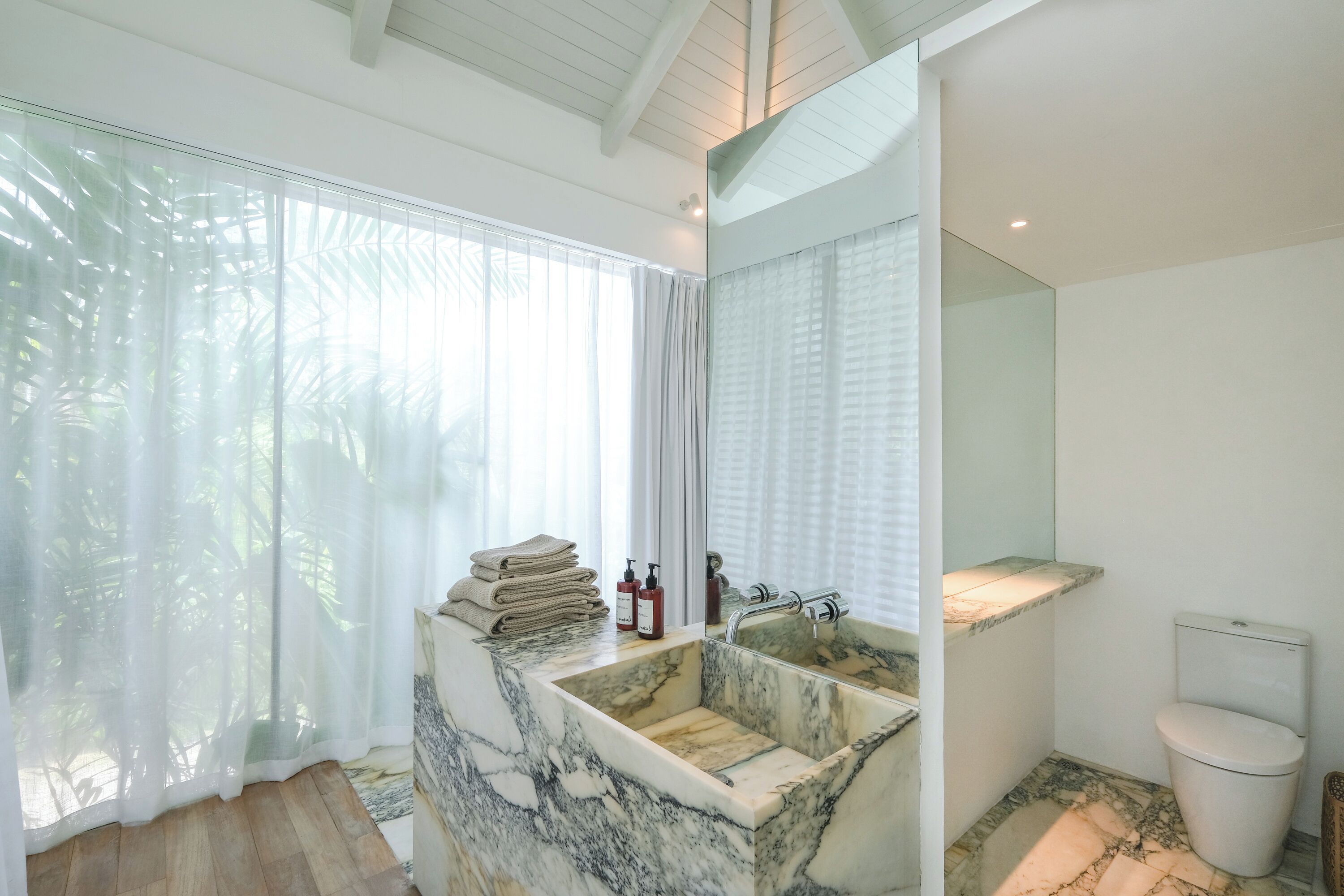 Une salle de bain moderne avec un lavabo en marbre, des toilettes et une grande fenêtre ornée de rideaux légers.