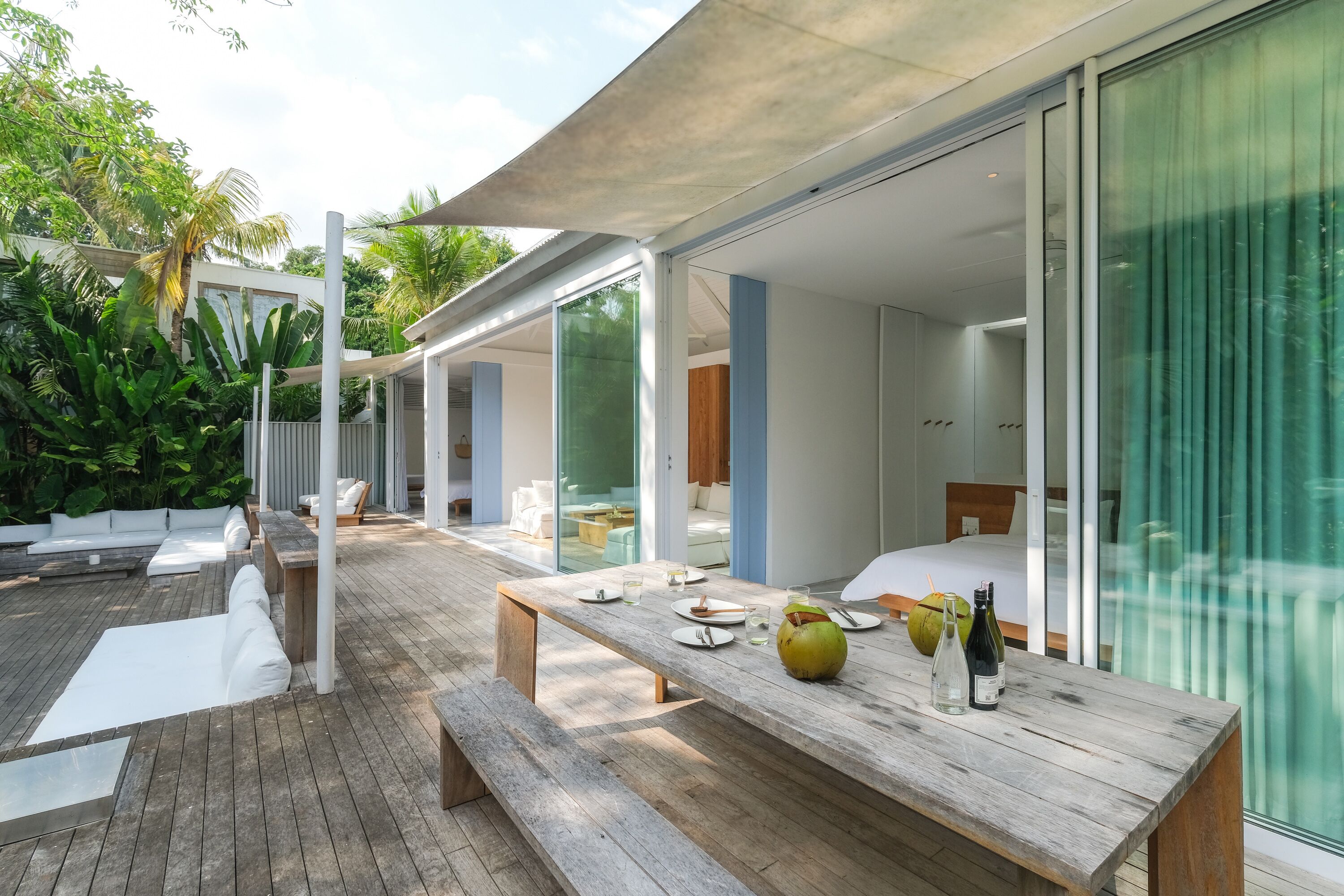 Een moderne eethoek buiten, met een houten tafel gedekt voor twee personen, omringd door een terras en een glazen deur die naar een slaapkamer leidt.