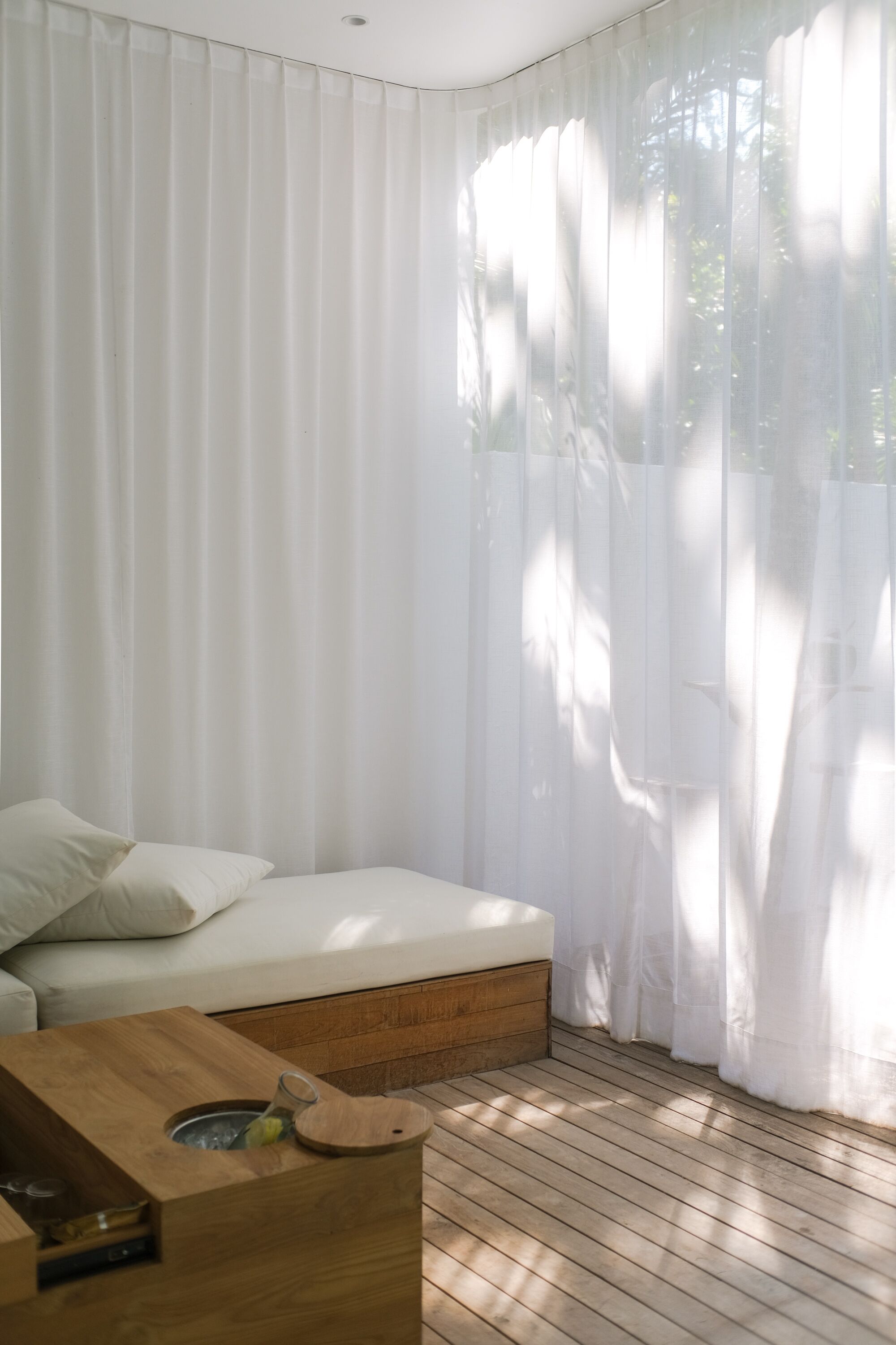 A room with a bed, a wooden side table with a glass, and sheer white curtains.